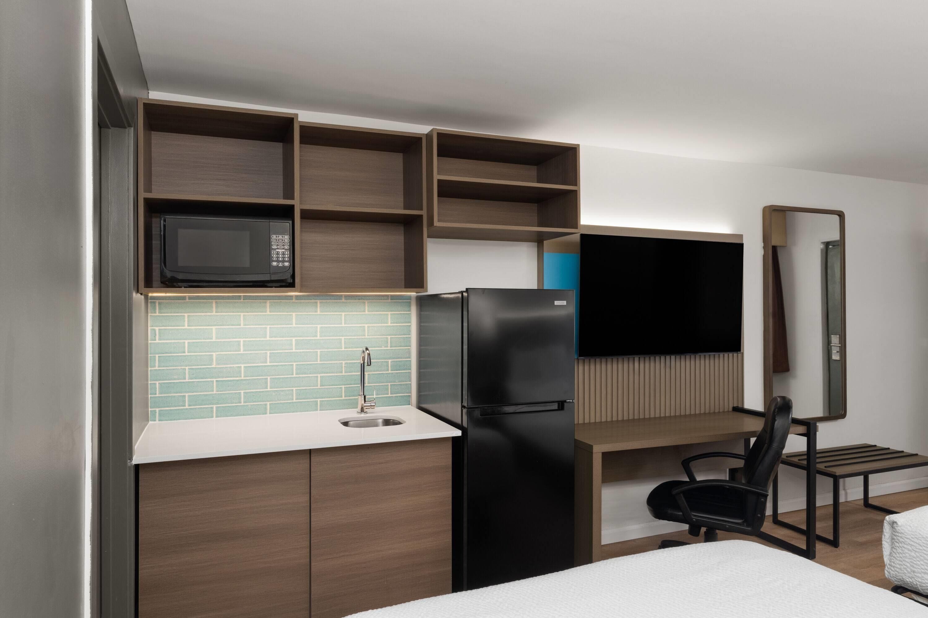 A compact hotel room with a kitchenette, a bed, a TV, and a mirror.