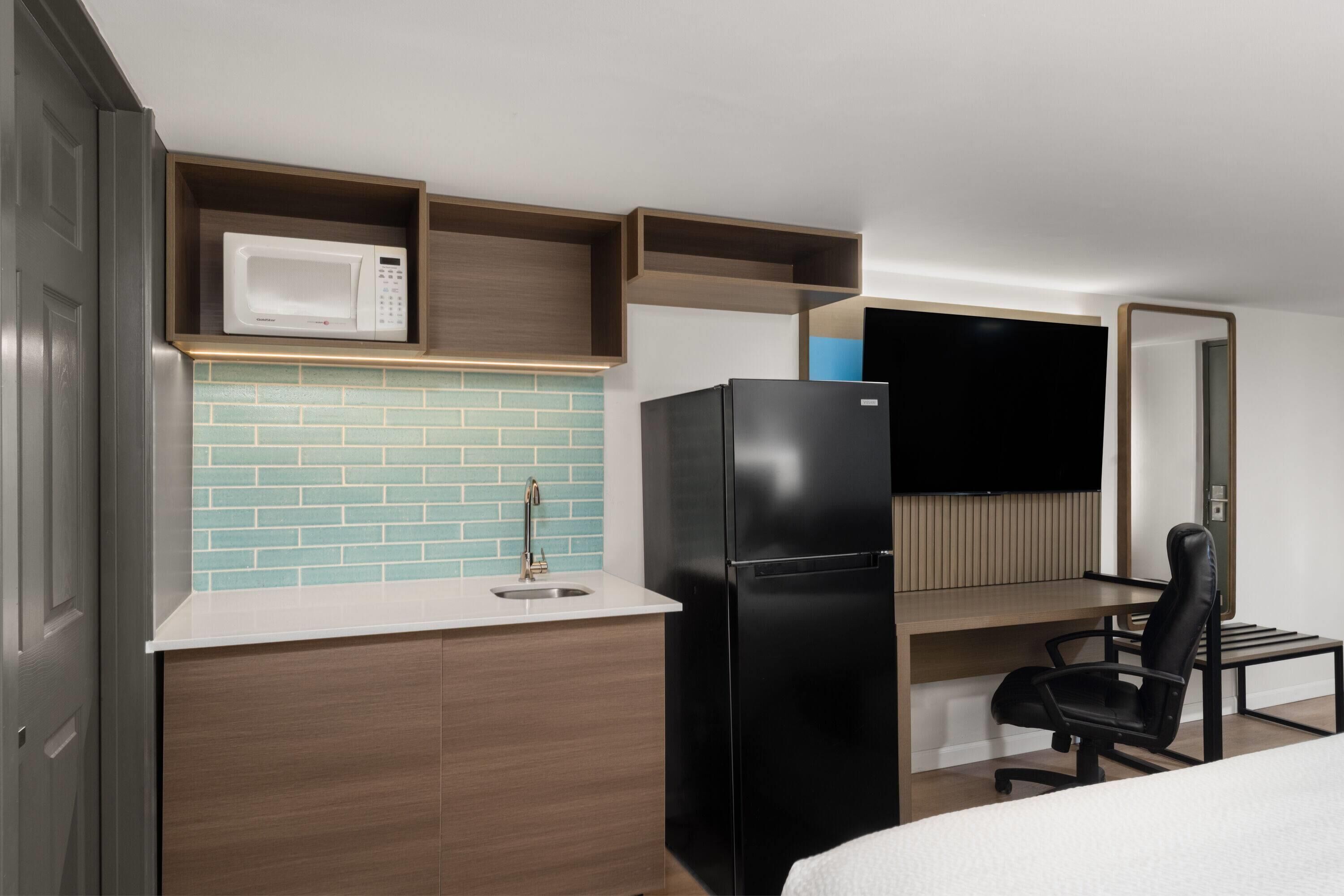 Eine kompakte Küche mit Mikrowelle, Kühlschrank und Spüle. An der Wand ist ein Fernseher angebracht und es gibt einen Stuhl im Essbereich.
