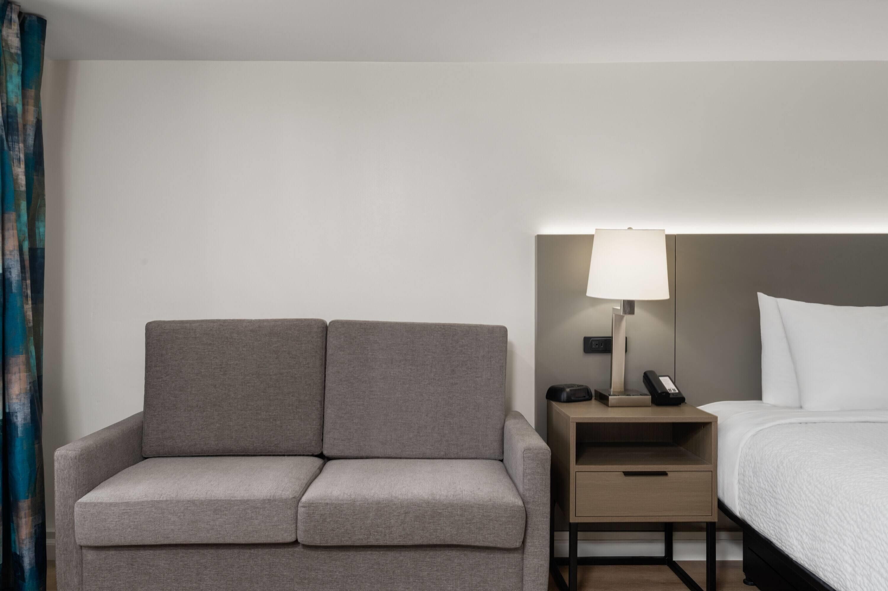 A hotel room with a grey sofa, a bedside table with a lamp, and a bed with white bedding.
