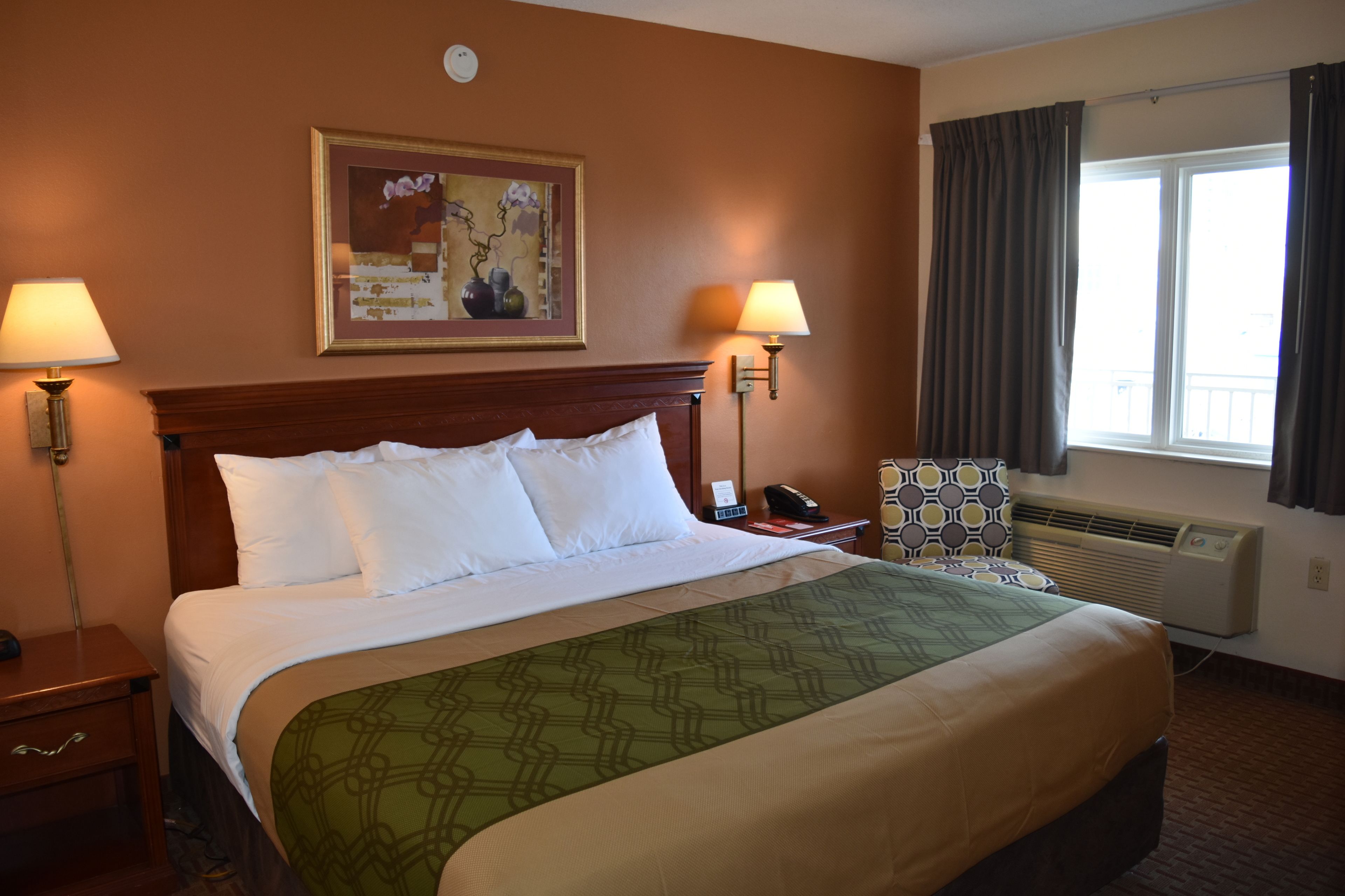 A hotel room with a large bed, two bedside lamps, a chair with a patterned cushion, a painting on the wall, and a window with curtains.