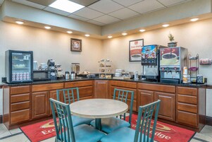 Una cocina de hotel con una mesa redonda y sillas, un refrigerador, máquinas de café y un microondas.