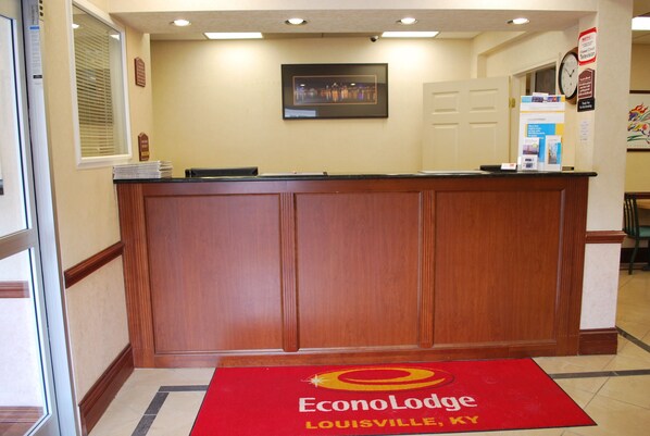 A reception desk with a framed picture above it, a clock on the wall, and a red carpet with the hotel name.