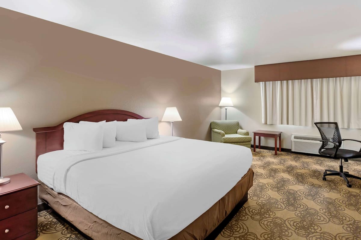 Quarto de hotel com uma cama grande, duas luminárias de cabeceira, uma poltrona verde, uma cadeira de escritório preta, uma mesa pequena e uma janela com cortinas.