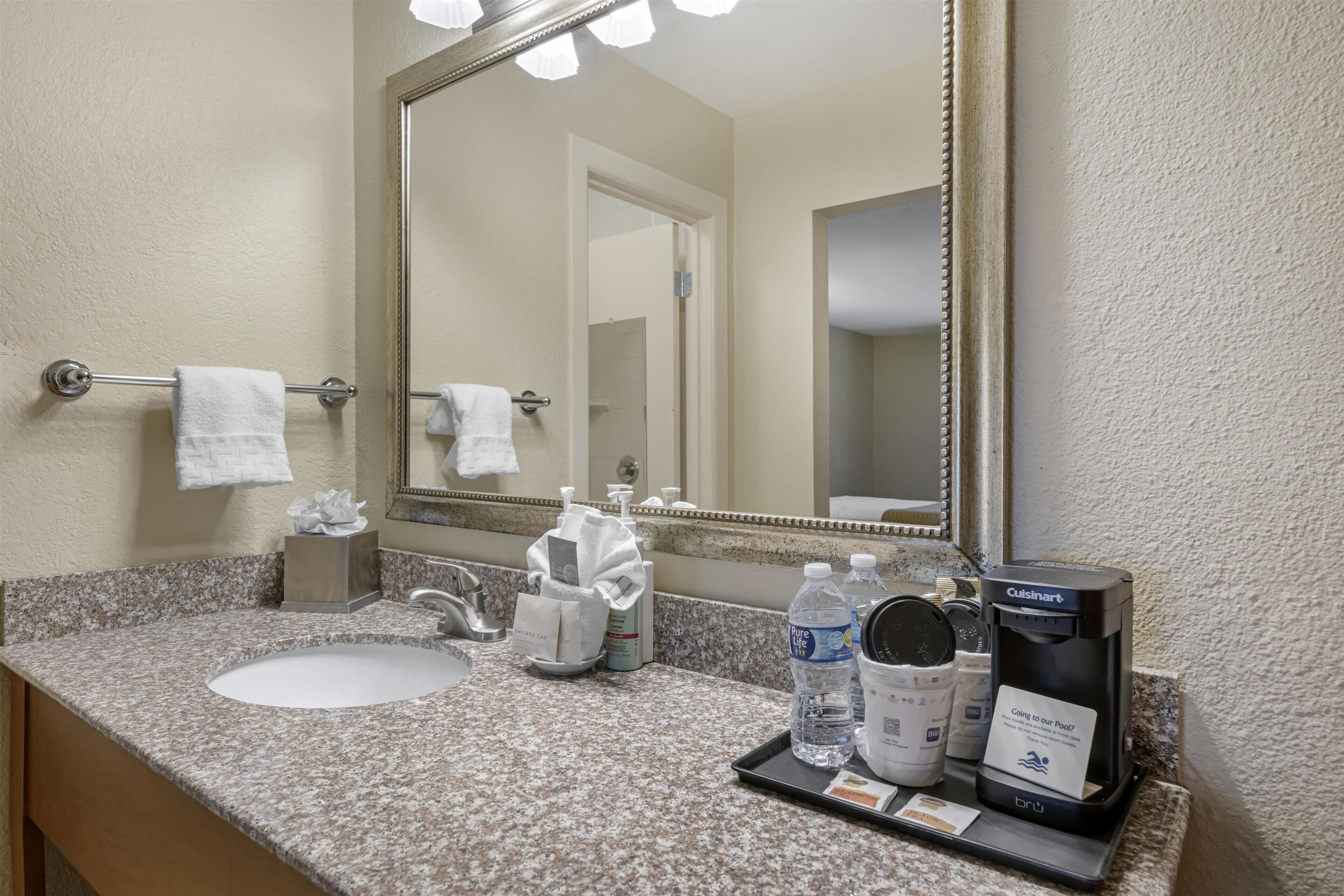 A hotel bathroom with a granite countertop, a white sink, a large mirror, and toiletries.