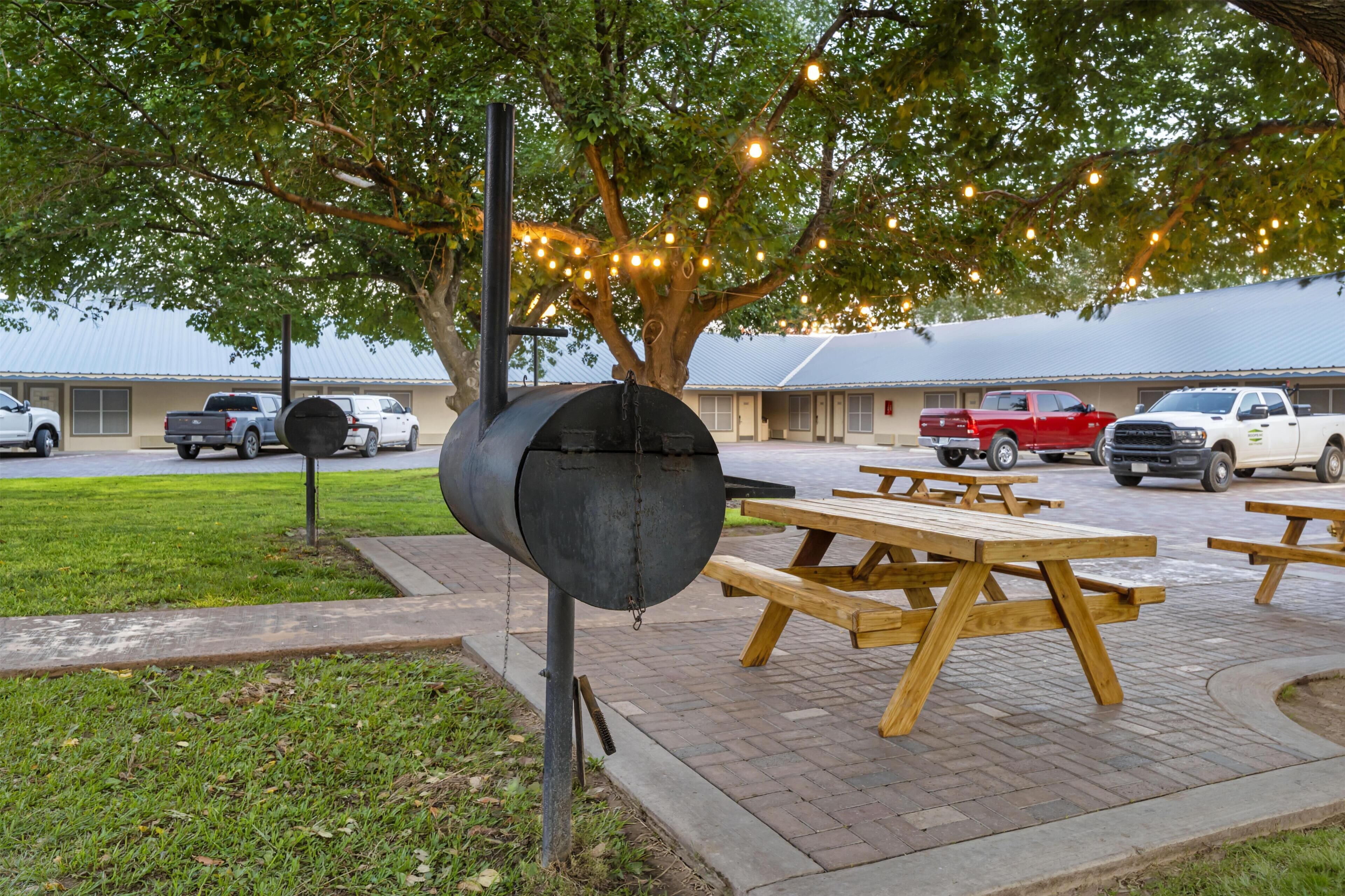A barbecue grill under a tree with picnic tables and parked cars in the background.