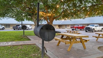 A barbecue grill under a tree with picnic tables and parked cars in the background.