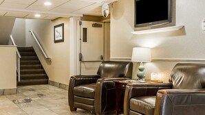 A hotel lobby with a flat-screen TV mounted on the wall, leather armchairs, a small table with a lamp, and a staircase leading to another area.