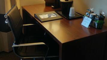A hotel room with a wooden desk, a black office chair, a lamp, a coffee maker, and a framed picture on the wall.