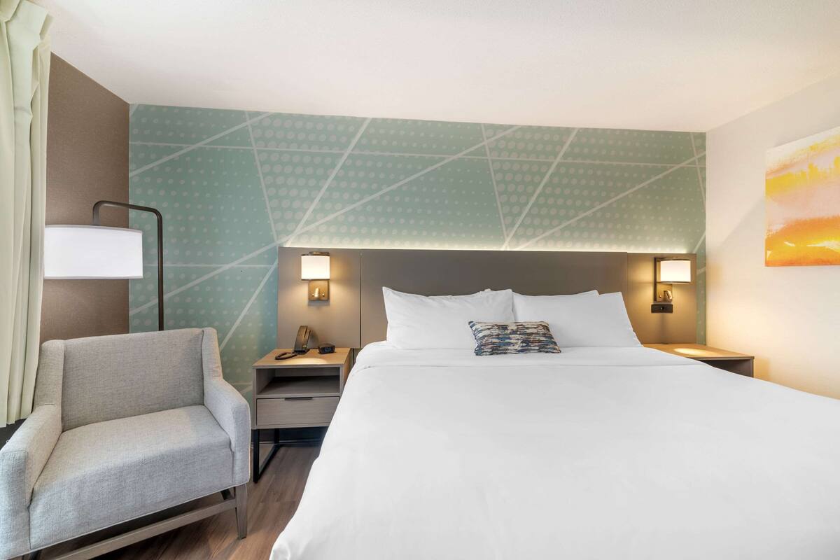 A hotel room with a large bed, a chair, a nightstand with a lamp, and a wall with a geometric pattern.