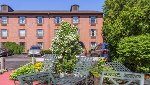 An outdoor seating area with metal chairs and a round table, surrounded by greenery and a brick building in the background.