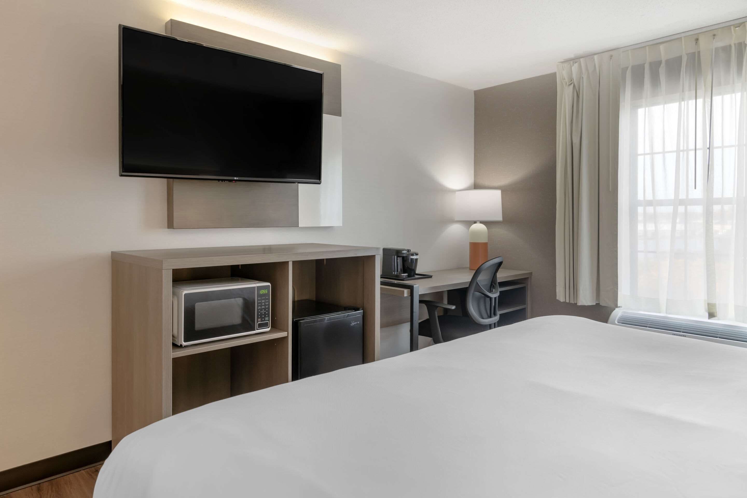 A hotel room with a large bed, a flat-screen TV mounted on the wall, a desk with a chair, a microwave, and a coffee maker.