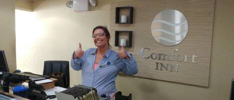 A woman giving a thumbs up in front of a Comfort INN sign, with a desk and computer visible in the background.
