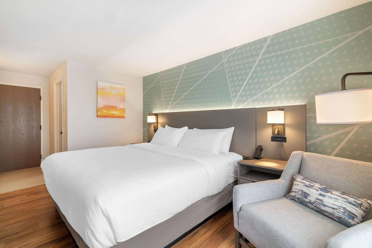 A hotel room with a large bed, a gray armchair, a nightstand with a lamp, and a wall with a geometric pattern.