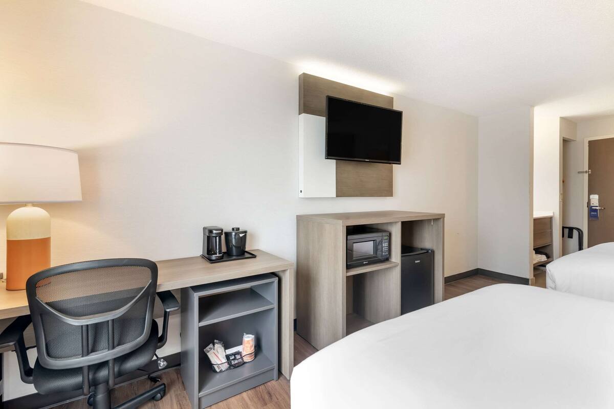A hotel room with a desk, a chair, a television mounted on the wall, a microwave, and a bed.