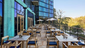 An outdoor dining area with wooden tables and chairs, a view of a modern building, and a railing overlooking a body of water. - Merriweather Lakehouse, Autograph Collection (Columbia)