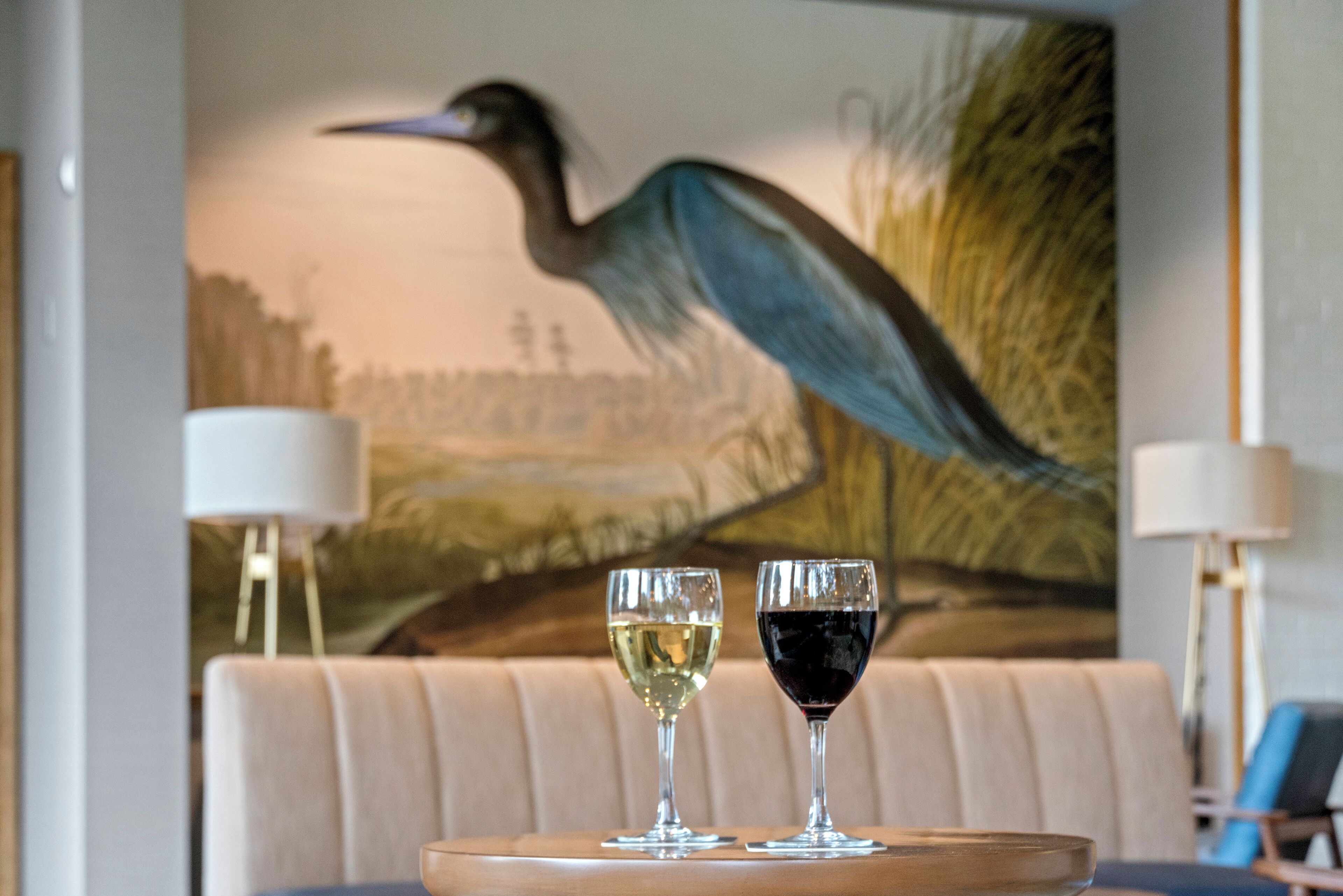 A dining table with two wine glasses, a wooden tray, and a painting of a bird in the background.