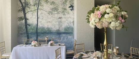 Une salle à manger où les tables sont dressées pour un événement formel, avec des nappes blanches, des chaises dorées et des centres de table floraux.