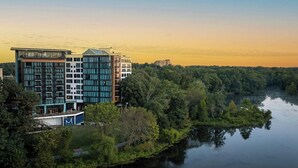 A modern hotel with multiple floors, large windows, and a view of a river and forested area. - Merriweather Lakehouse, Autograph Collection (Columbia)