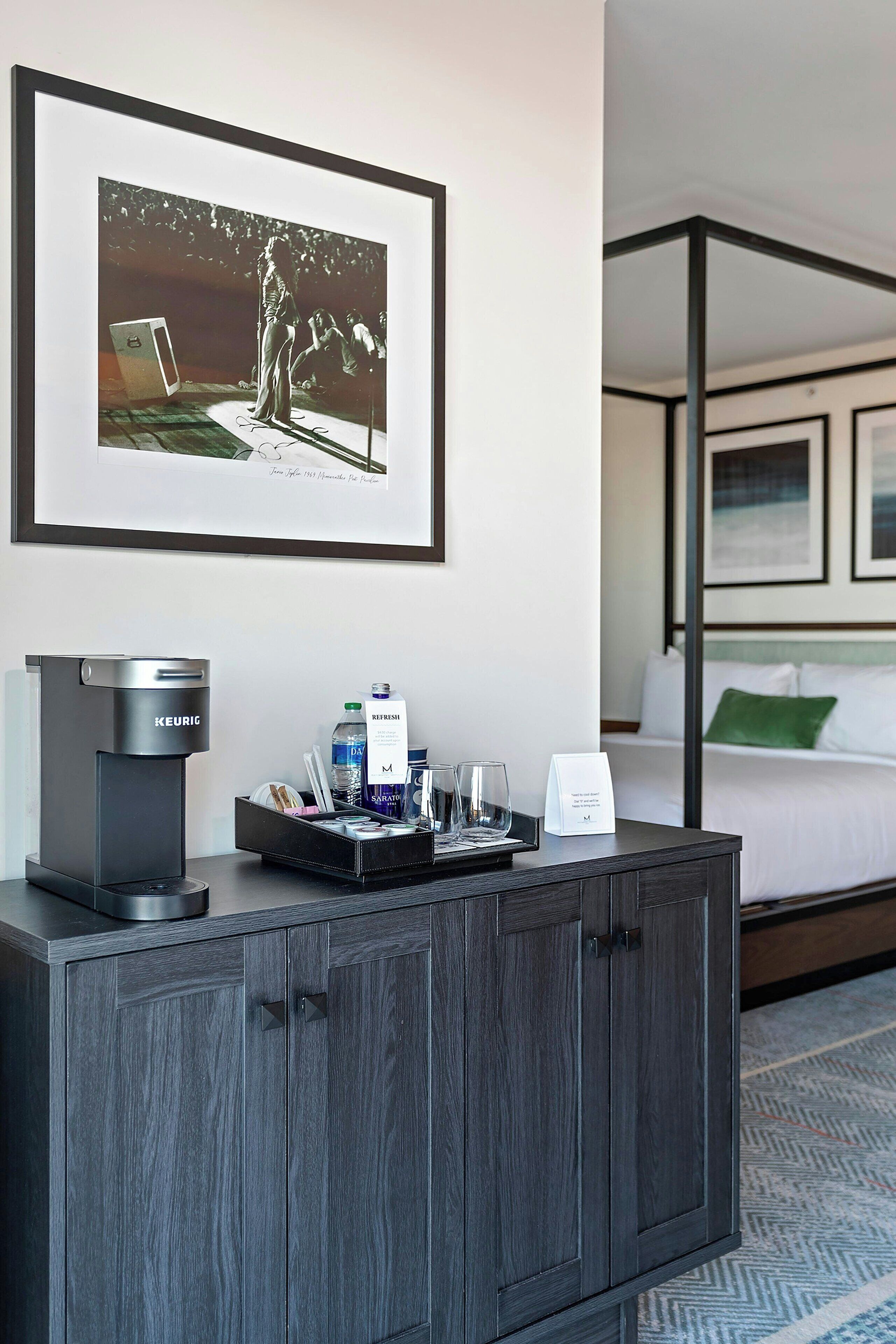 Una habitación de hotel con un minibár, un cuadro en la pared y una cama con un almohadón verde.