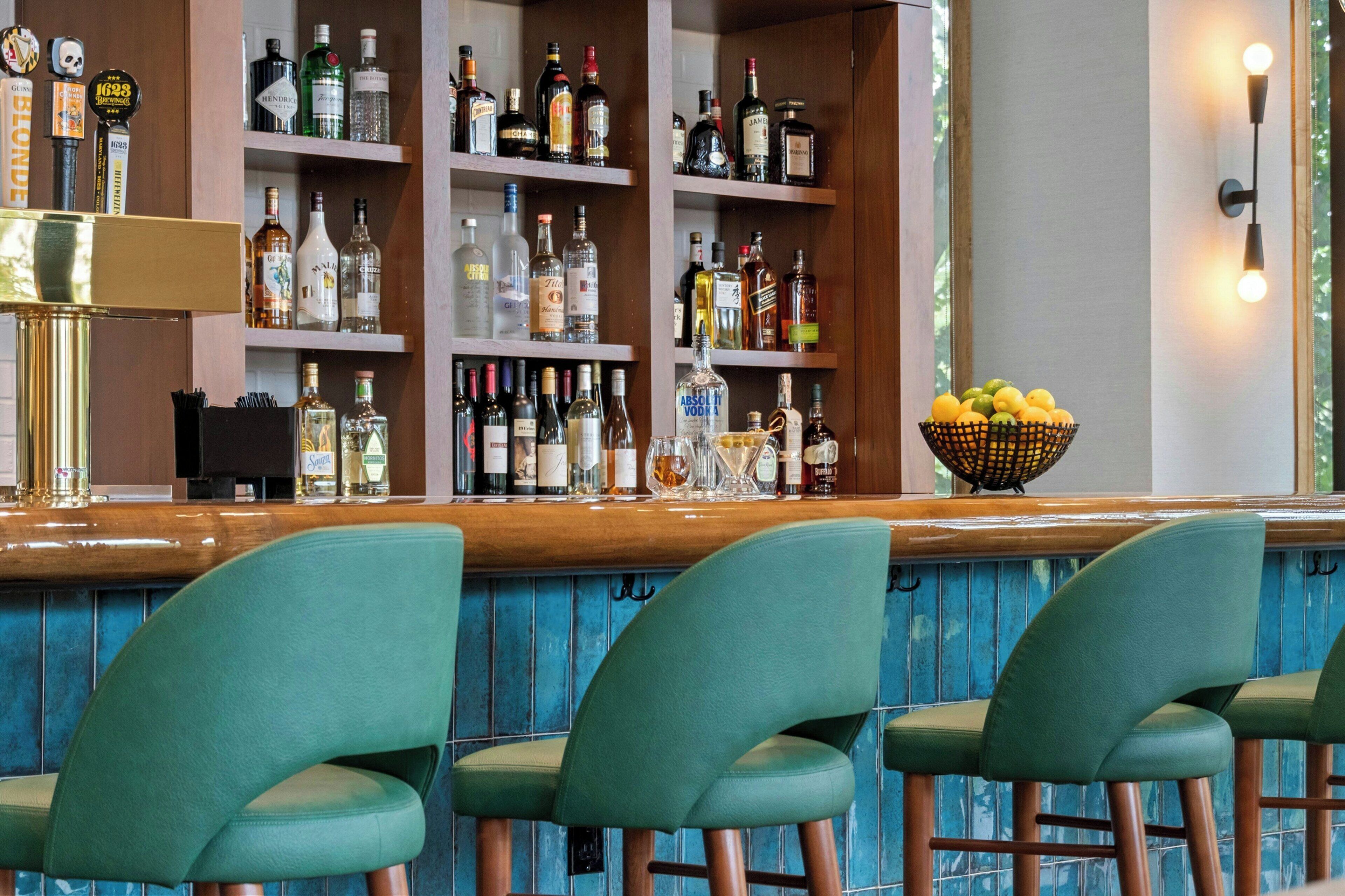 Zona de bar con taburetes verdes, estantes llenos de botellas de licor y un frutero sobre la barra.