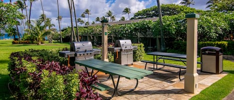 Un patio al aire libre con una parrilla, una mesa de picnic y bancos, rodeado de abundante vegetación y palmeras.