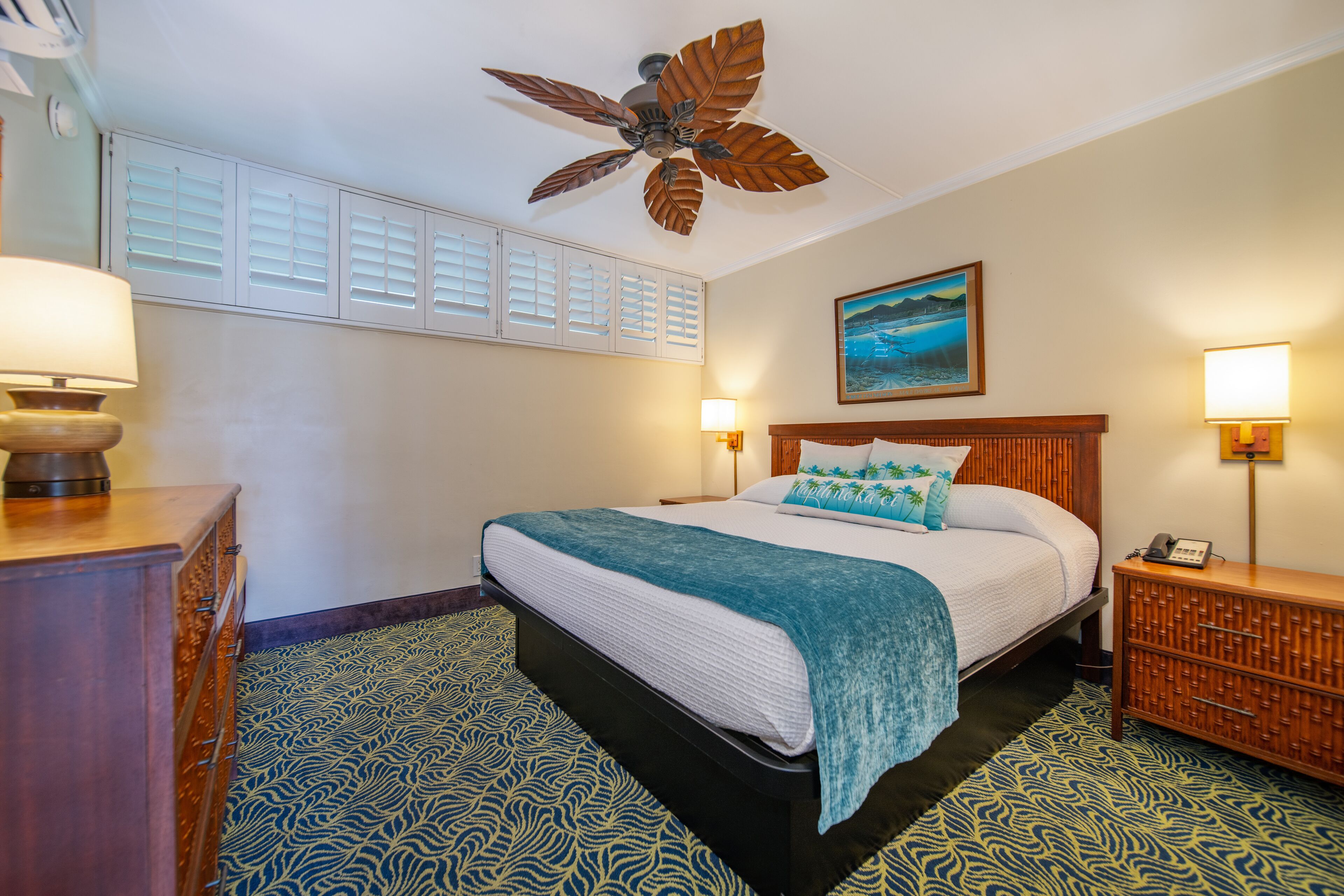A hotel room with a bed, a ceiling fan, a nightstand, a lamp, and a painting on the wall.