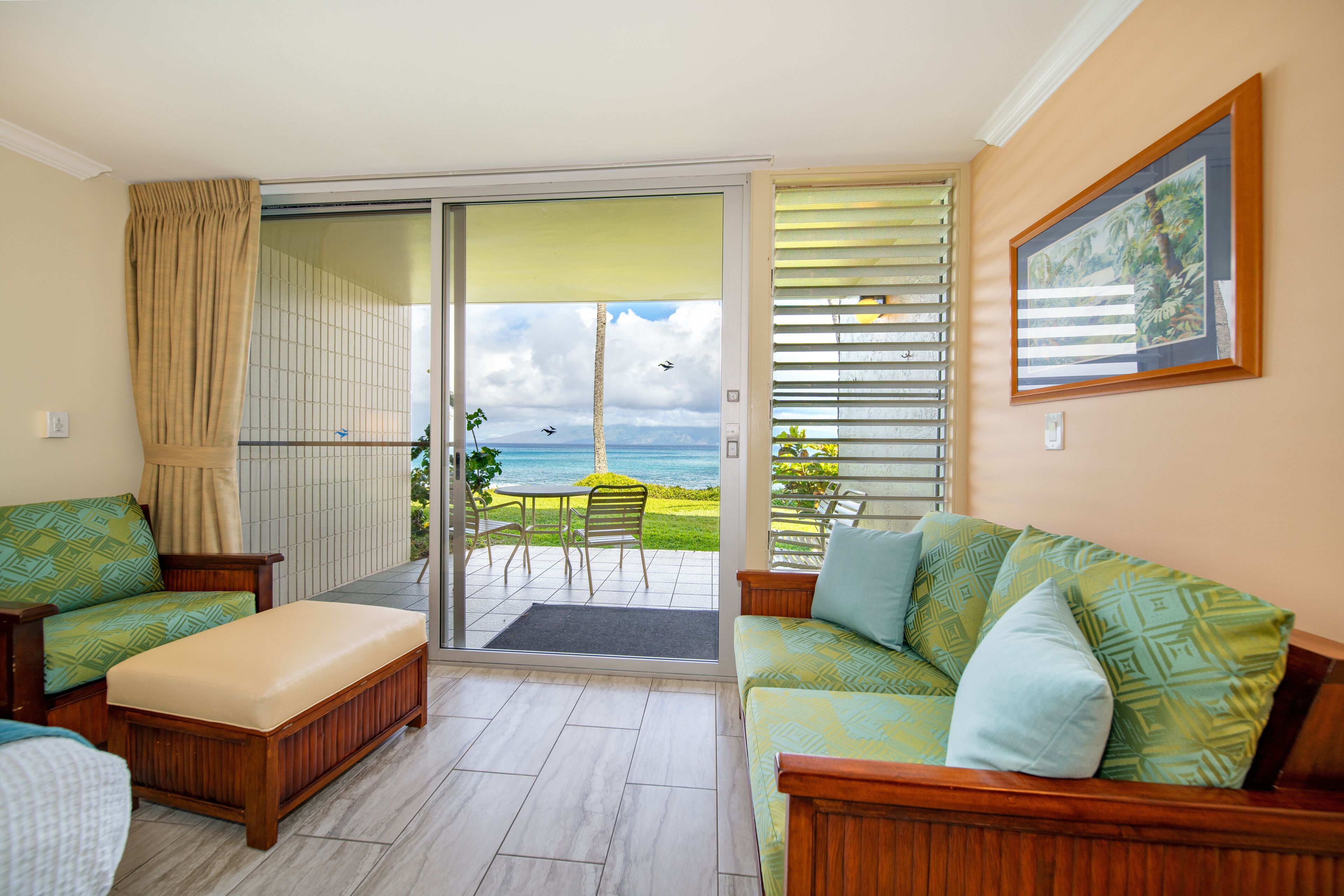 A room with a sliding glass door leading to a balcony with a view of the ocean and a mountain. There are two sofas and a bench inside the room.