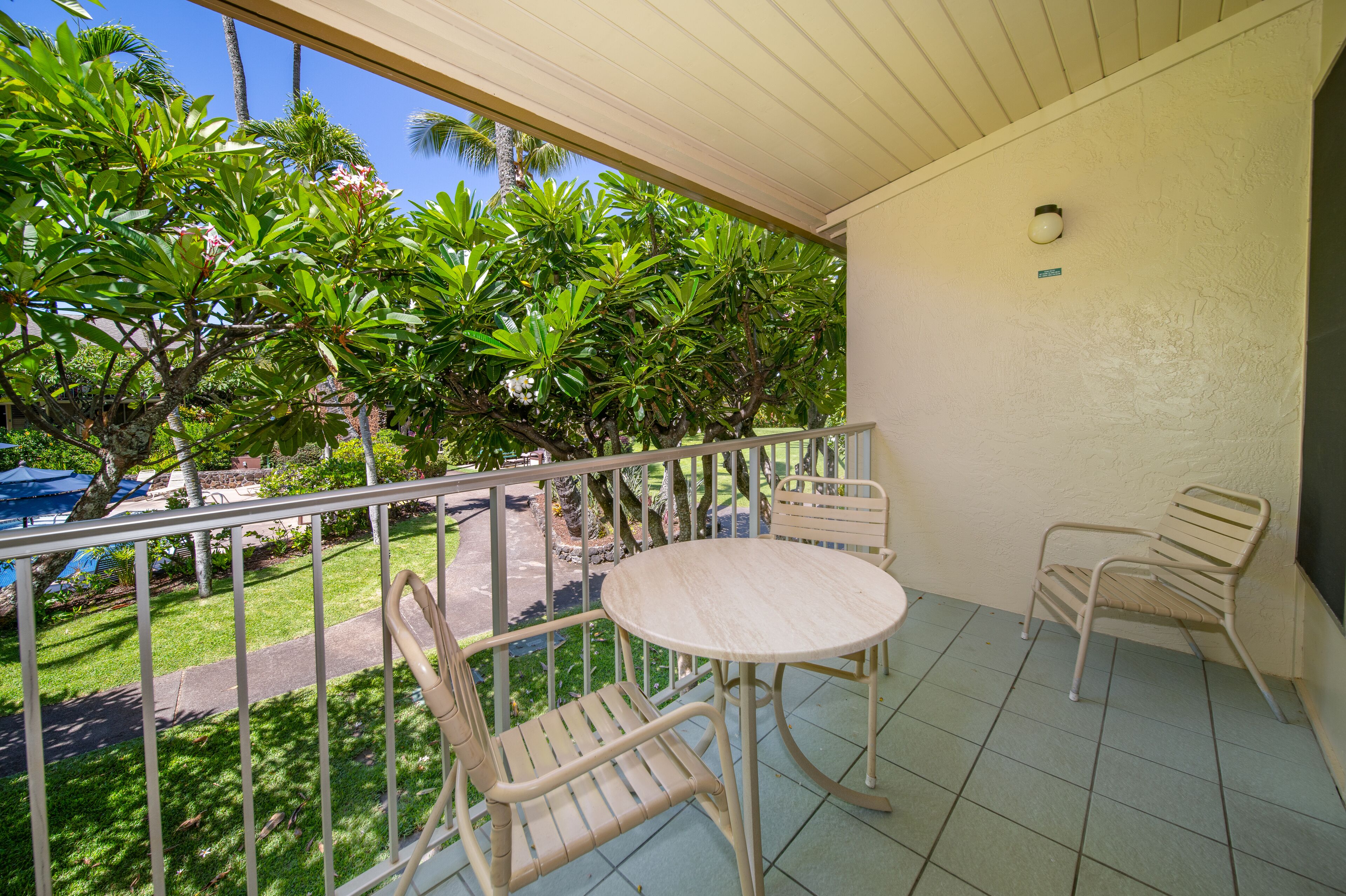 A balcony with a round table and two chairs, surrounded by lush greenery and a clear blue sky.