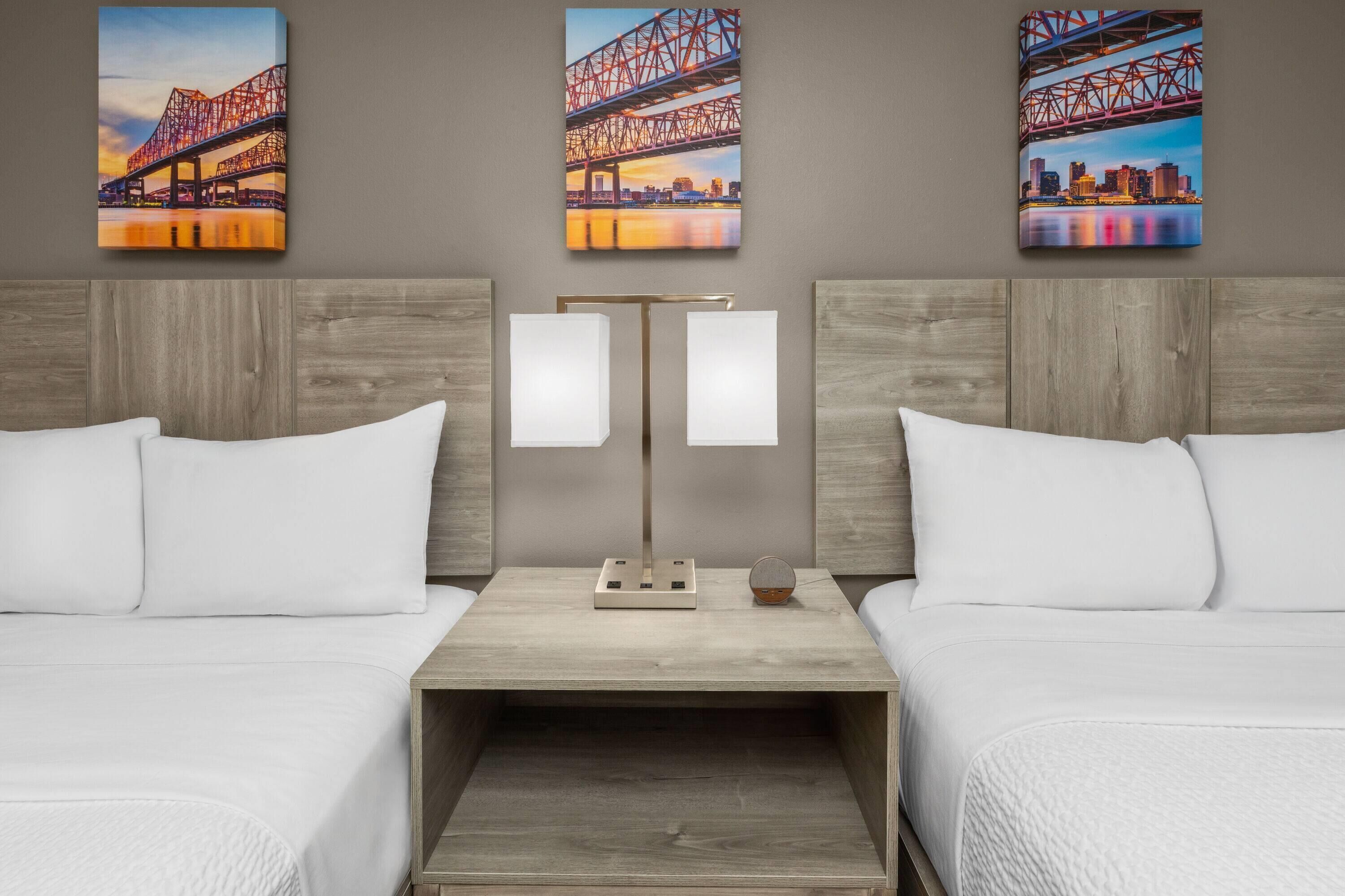 A hotel room with two beds, a nightstand with a lamp, and three paintings of a bridge on the wall.