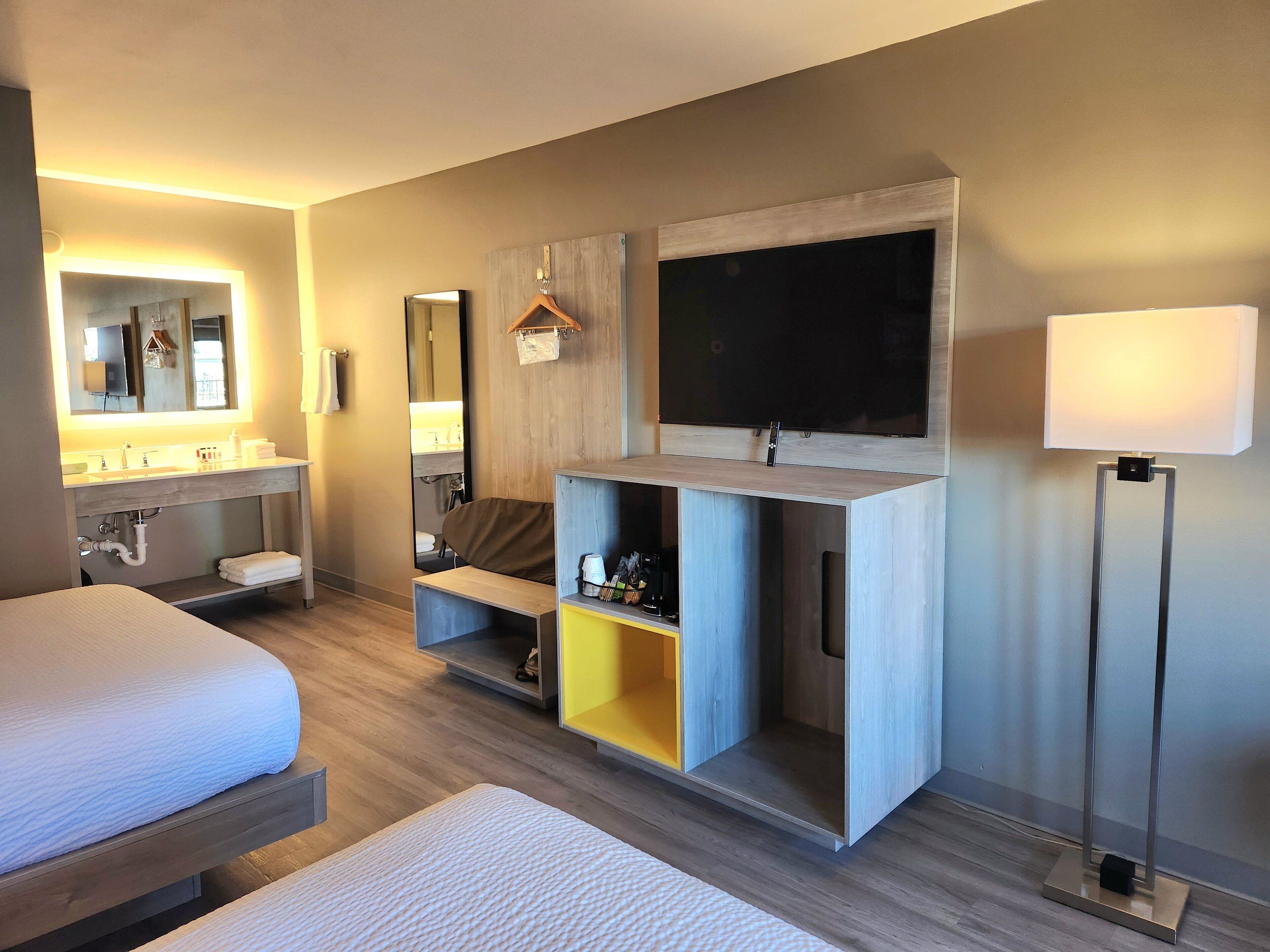 A modern hotel room with a bed, a vanity with a mirror, a flat-screen TV mounted on a wooden cabinet, and a bedside lamp.