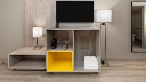 Ein modernes Hotelzimmer mit Flachbildfernseher an einer Holzvertäfelung, einem Nachttisch mit Lampe, einer Kaffeemaschine und einer gelben Aufbewahrungsbox.