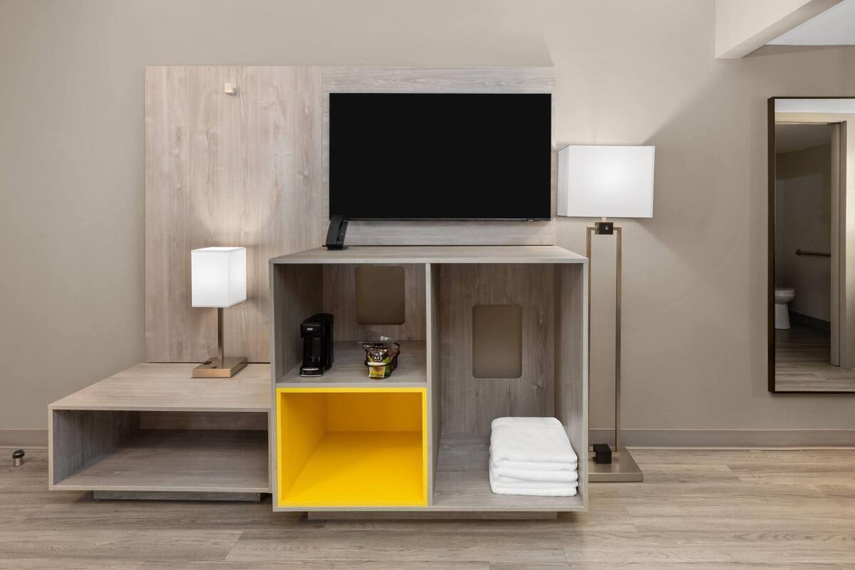 A modern hotel room with a flat-screen TV mounted on a wooden panel, a bedside table with a lamp, a coffee maker, and a yellow storage box.