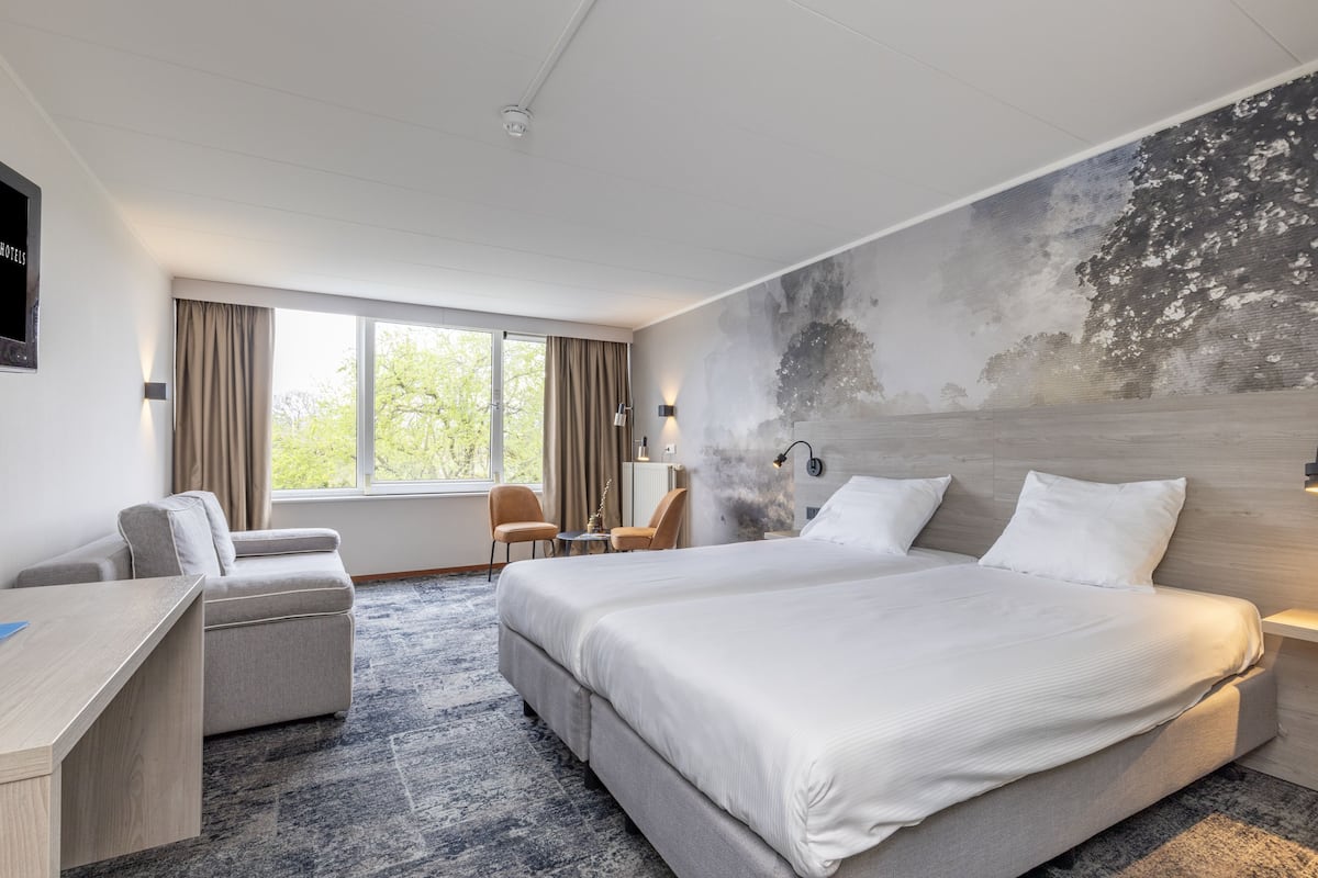 Ein modernes Hotelzimmer mit einem großen Bett, einem Schreibtisch, einem Sessel und Blick ins Freie.