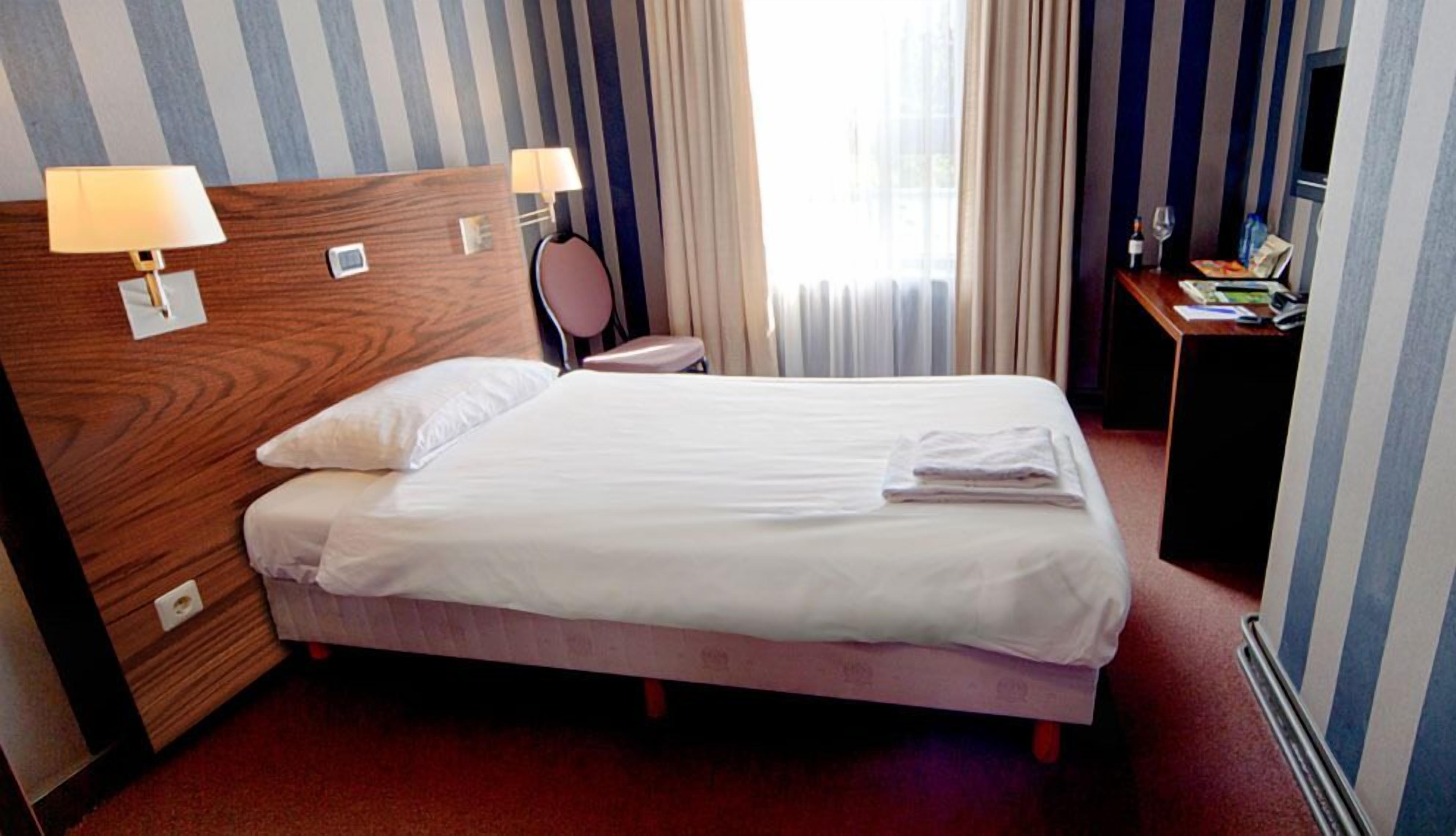 Ein Hotelzimmer mit einem Bett, zwei Nachttischlampen, einem kleinen Tisch mit Flasche und Gläsern sowie einem Fenster mit Vorhängen.