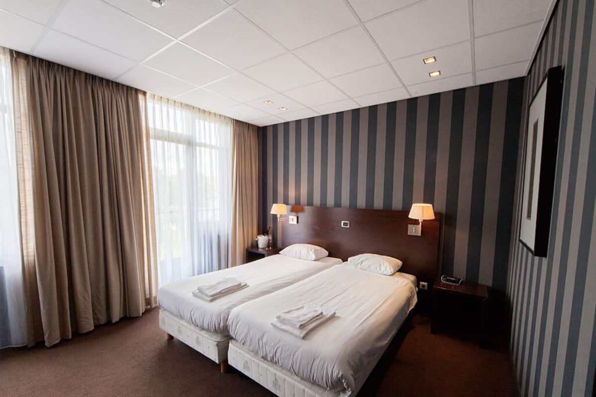 Ein Hotelzimmer mit zwei Betten, einem großen Fenster mit Vorhängen, einer gestreiften Wand und einem an der Wand befestigten Fernseher.