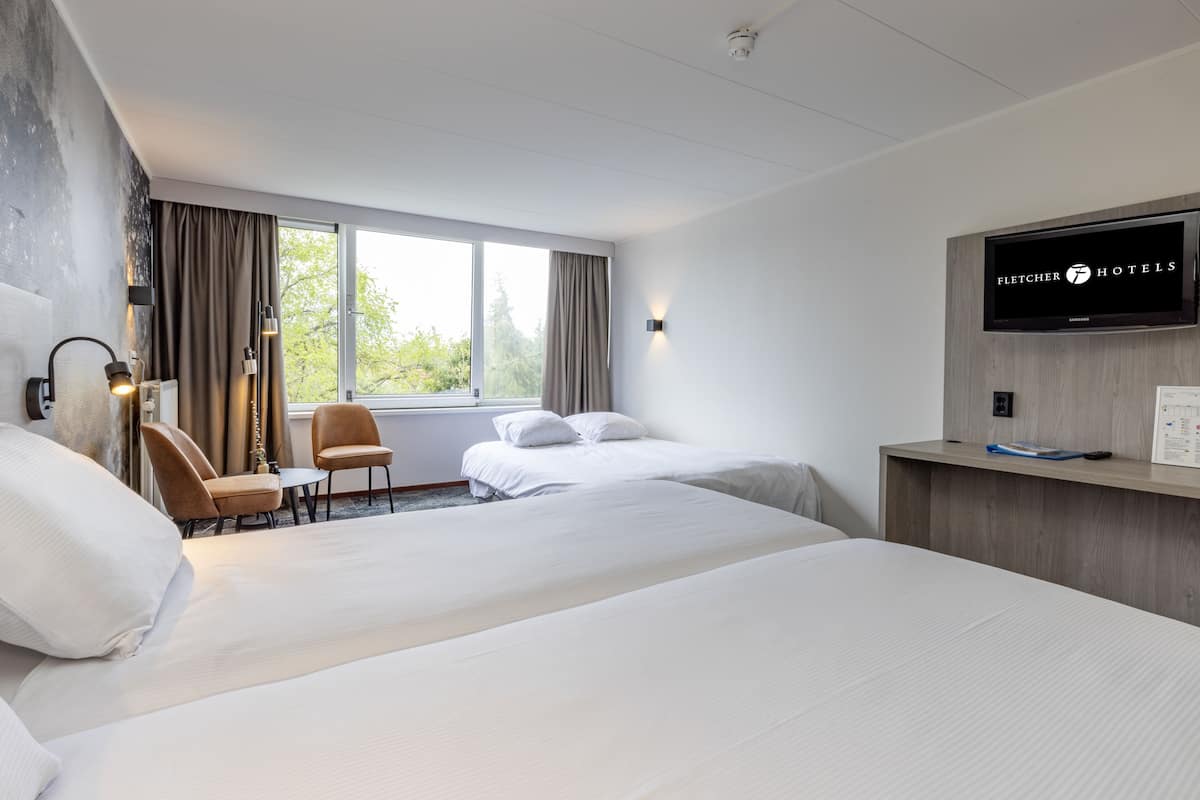 Ein Hotelzimmer mit zwei Betten, einem Schreibtisch, einem Sessel, einem Fernseher und einem Fenster mit Vorhängen.