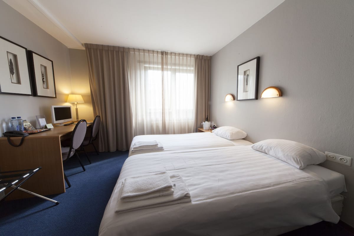 Ein Hotelzimmer mit zwei Betten, einem Schreibtisch mit Computer, einem Stuhl, einer Schreibtischlampe und einem Fenster mit Vorhängen.
