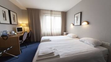 A hotel room with two beds, a desk with a computer, a chair, a desk lamp, and a window with curtains.