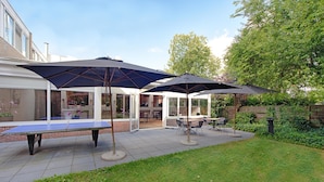 En udendørs patio med pingpongbord, to parasoller og siddepladser.