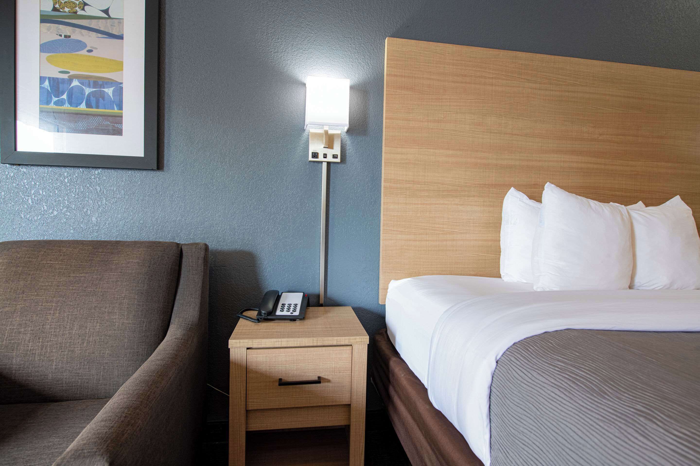 A hotel room with a bed, a nightstand with a phone, a lamp, and a framed picture on the wall.