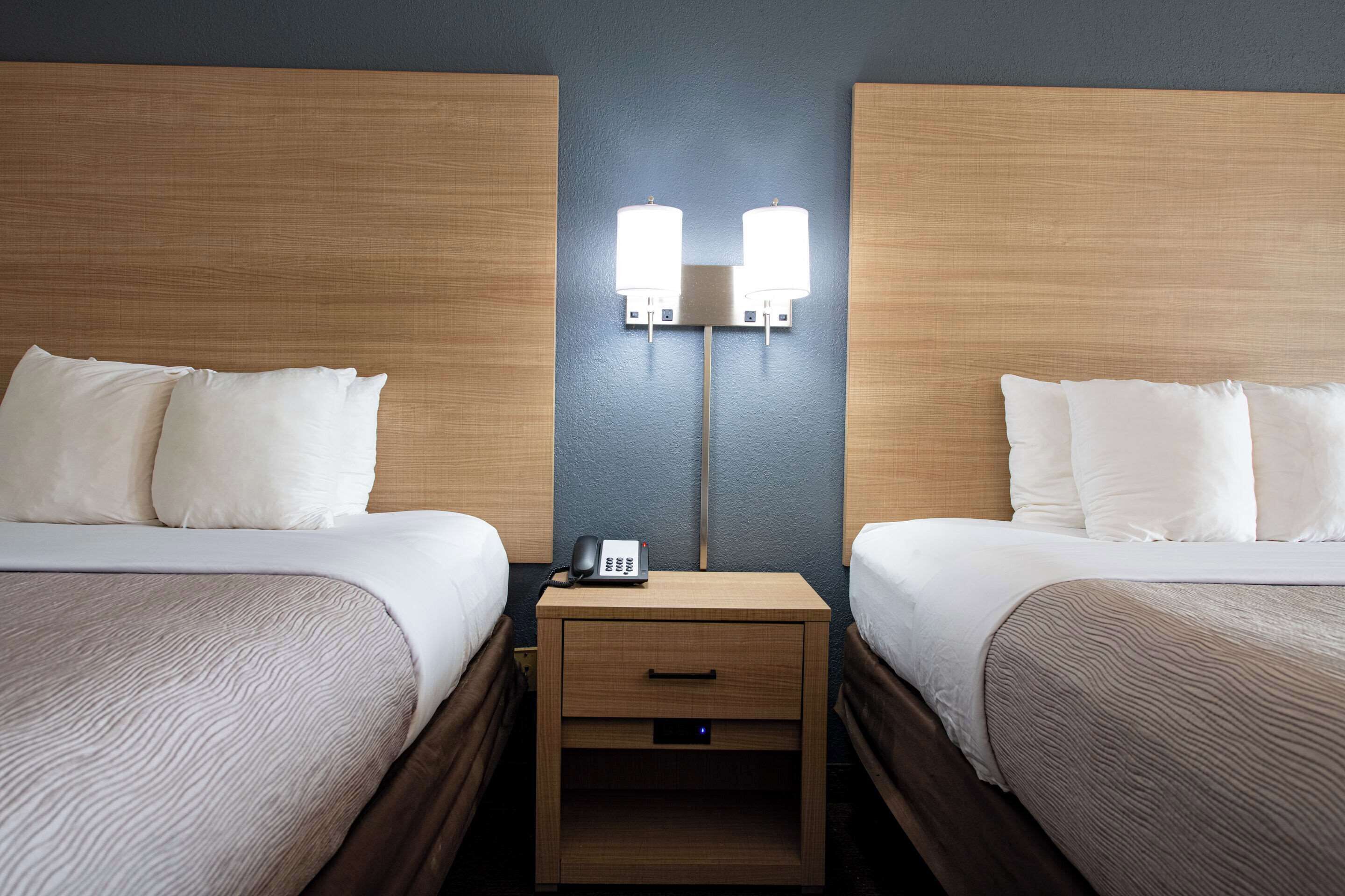 A hotel room with two beds, a nightstand with a phone, and wall-mounted lights.