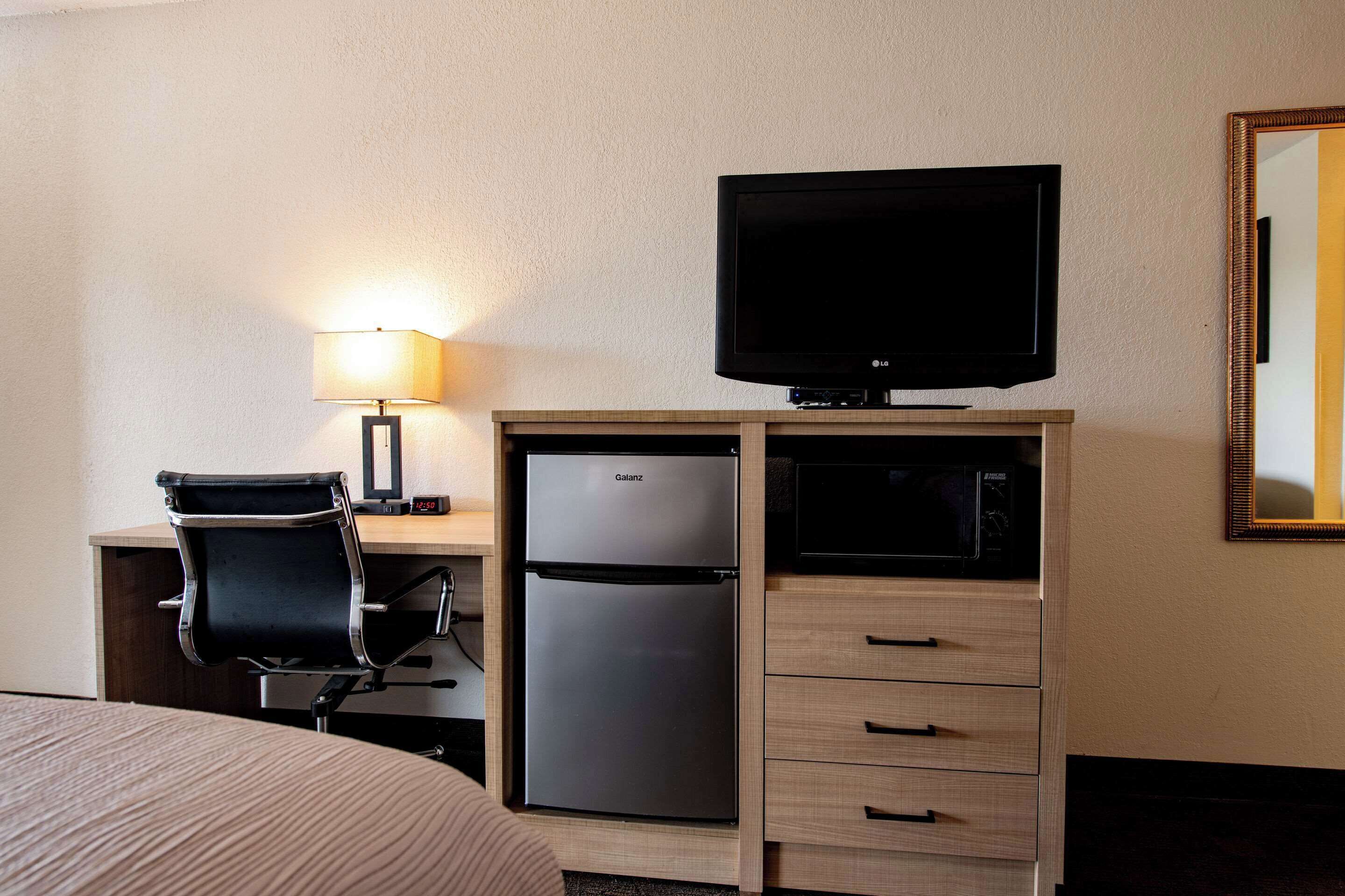 A hotel room with a bed, a desk with a computer, a refrigerator, a television, a lamp, and a mirror.