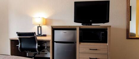 A hotel room with a bed, a desk with a computer, a refrigerator, a television, a lamp, and a mirror.