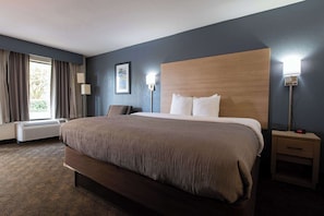A hotel room with a large bed, a bedside table with a lamp, a window with curtains, and a wall-mounted air conditioner.