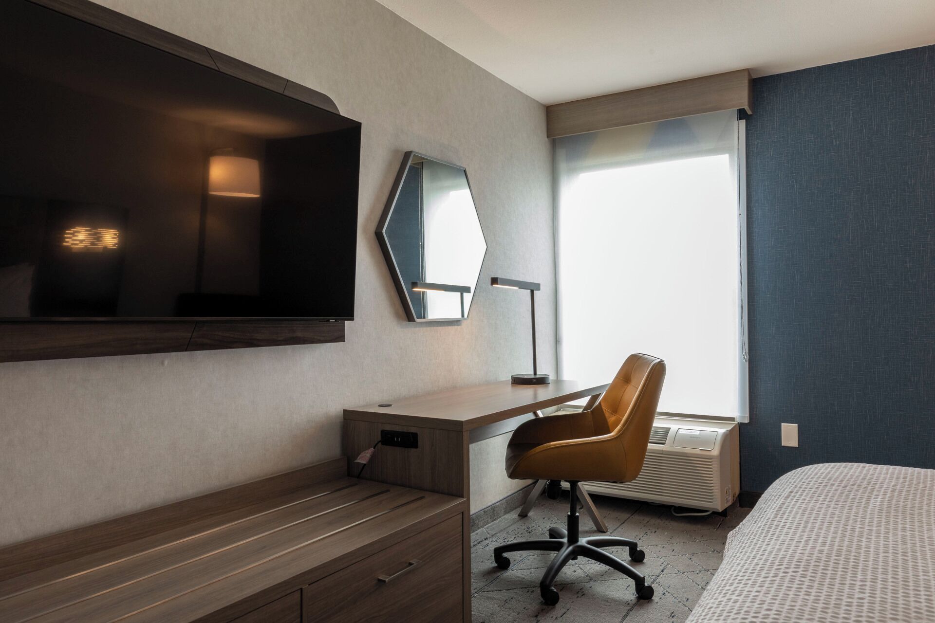 Camera d'albergo con una grande TV a schermo piatto fissata al muro, una scrivania con una sedia, un letto e un'ampia finestra con tende.
