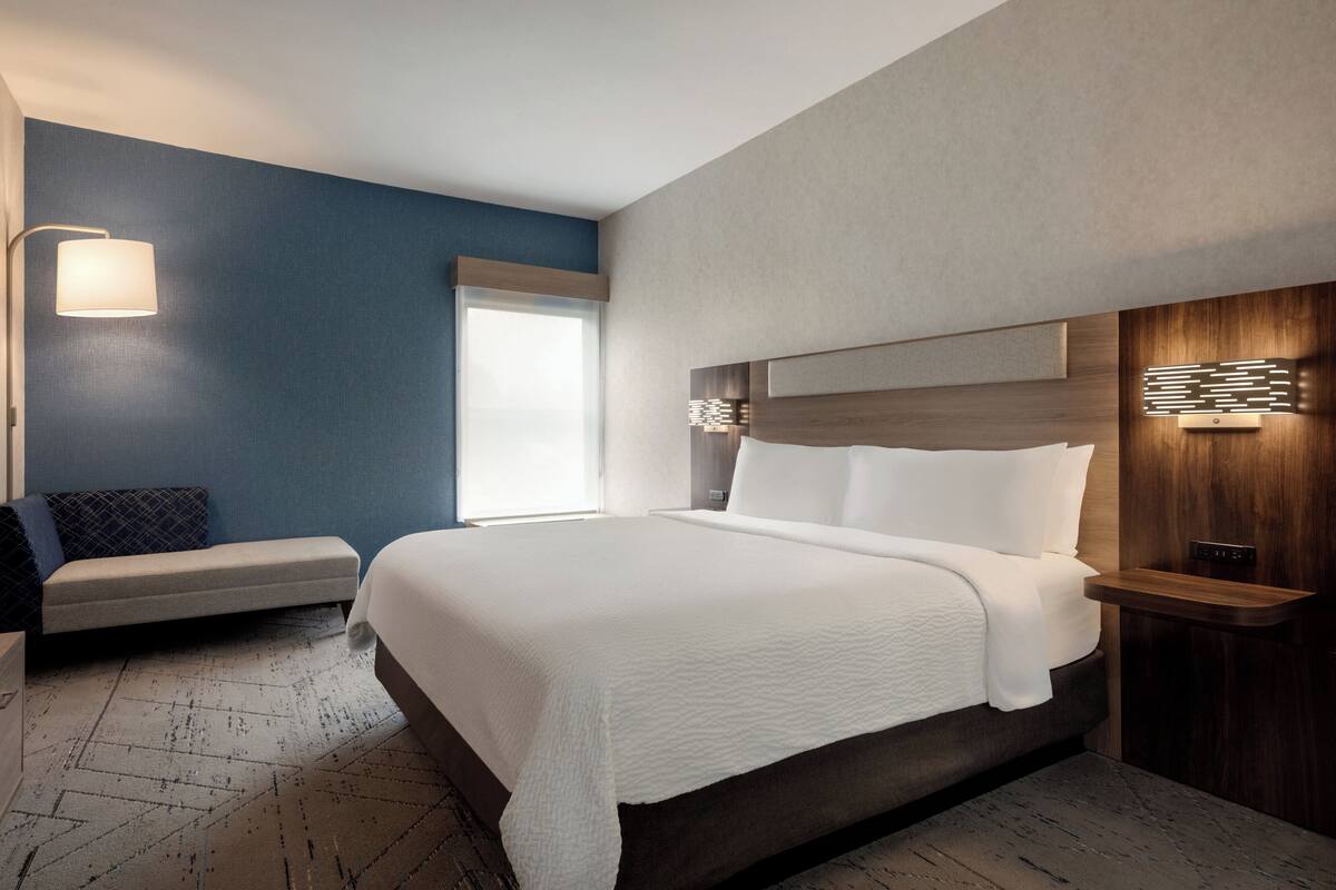 A modern hotel room with a large bed, a bedside table, a wall-mounted lamp, and a bench with a cushion.