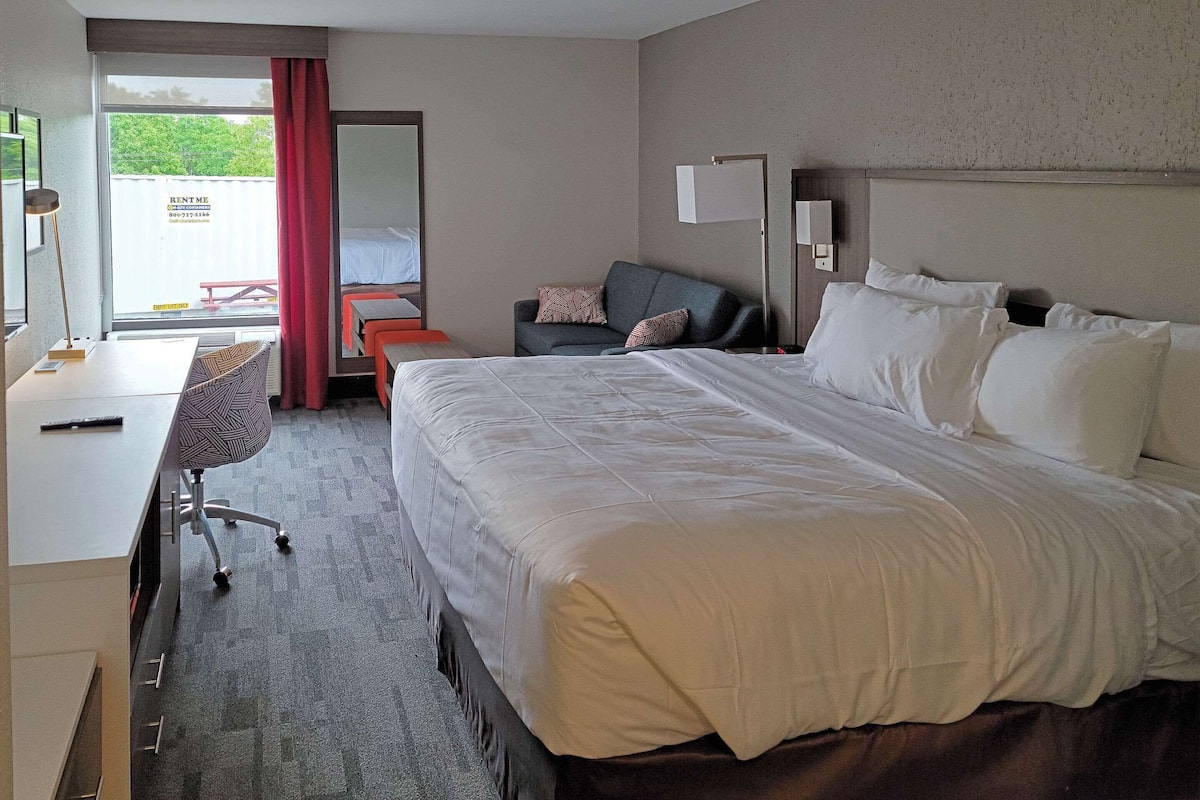 A hotel room with a large bed, a desk, a chair, a sofa, and a window with a view of a red building.