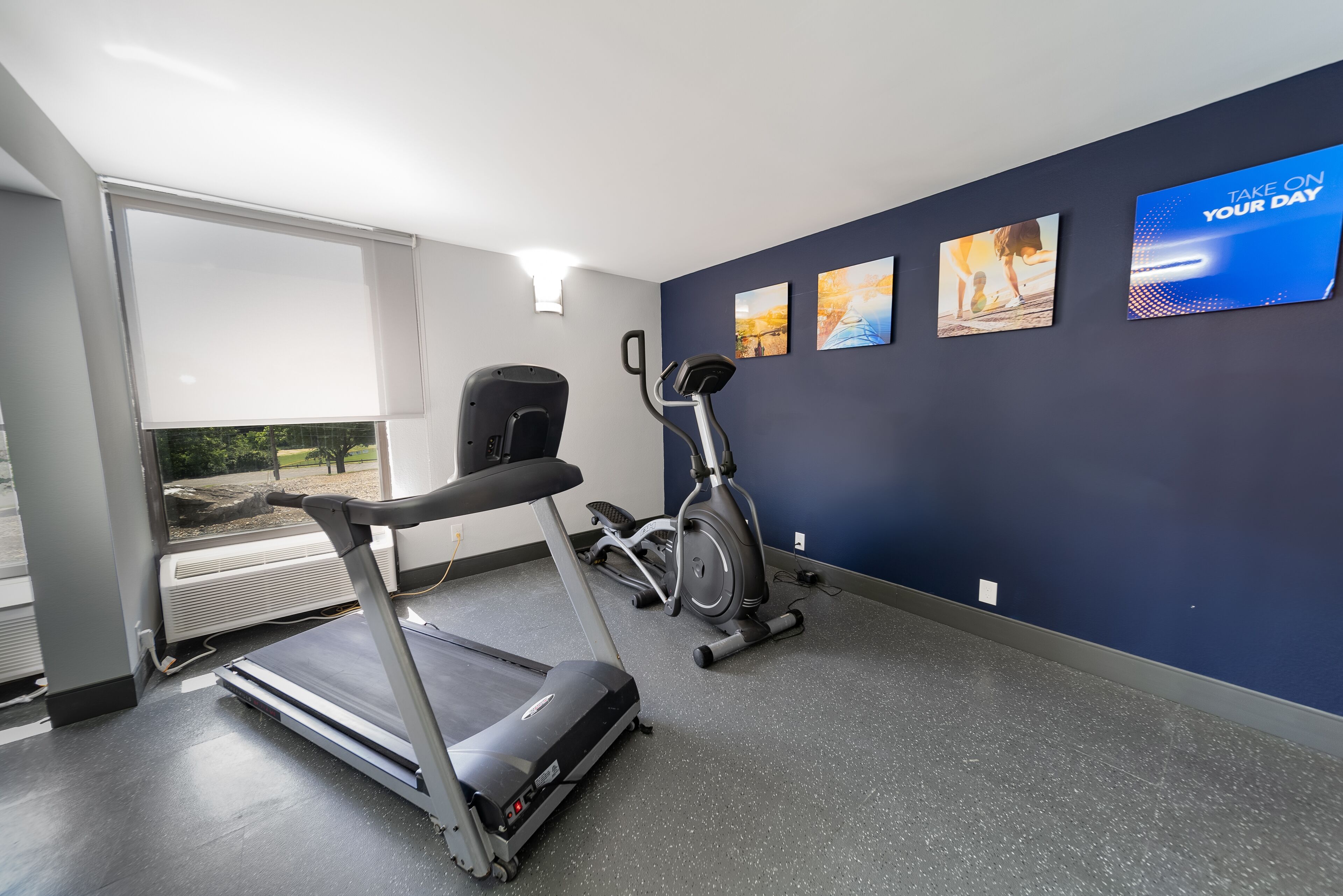 Een sportschool met een loopband, elliptische trainer en hometrainer, grote ramen en motiverende muurkunst.