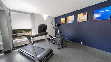 Een sportschool met een loopband, elliptische trainer en hometrainer, grote ramen en motiverende muurkunst.