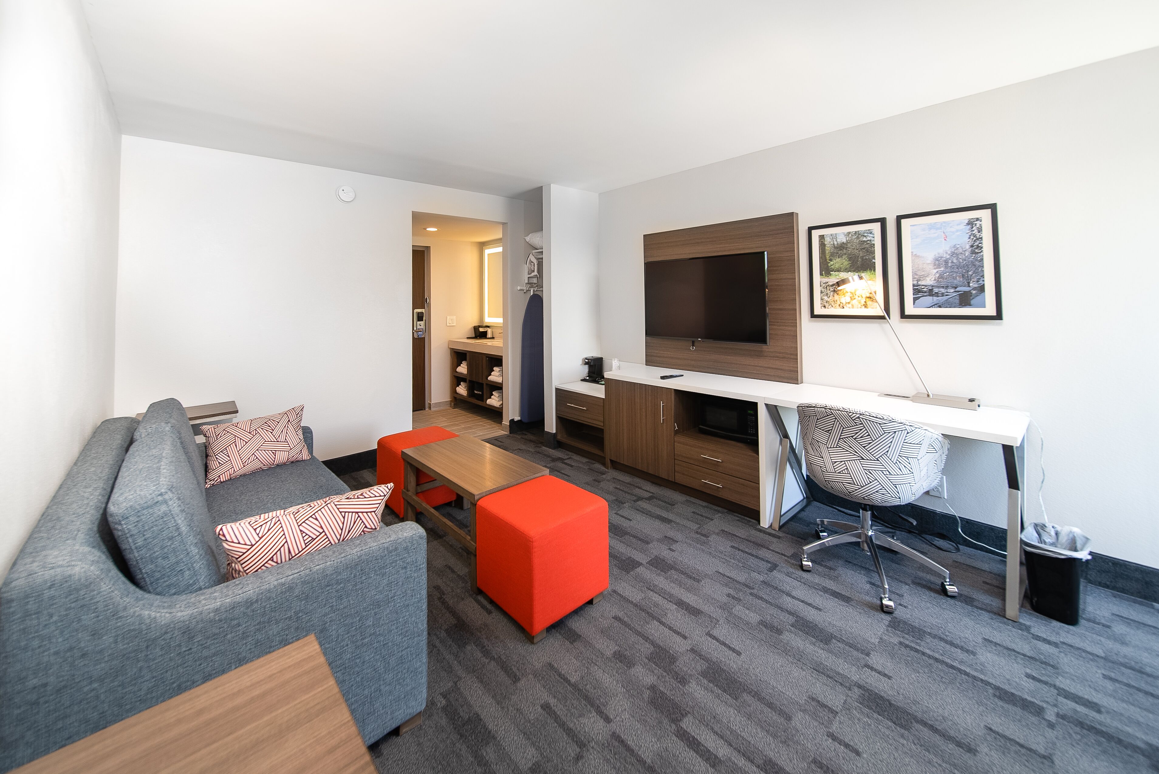 A modern living room with a grey sofa, red ottoman, and a wooden coffee table. A desk with a chair and a TV is against the wall.
